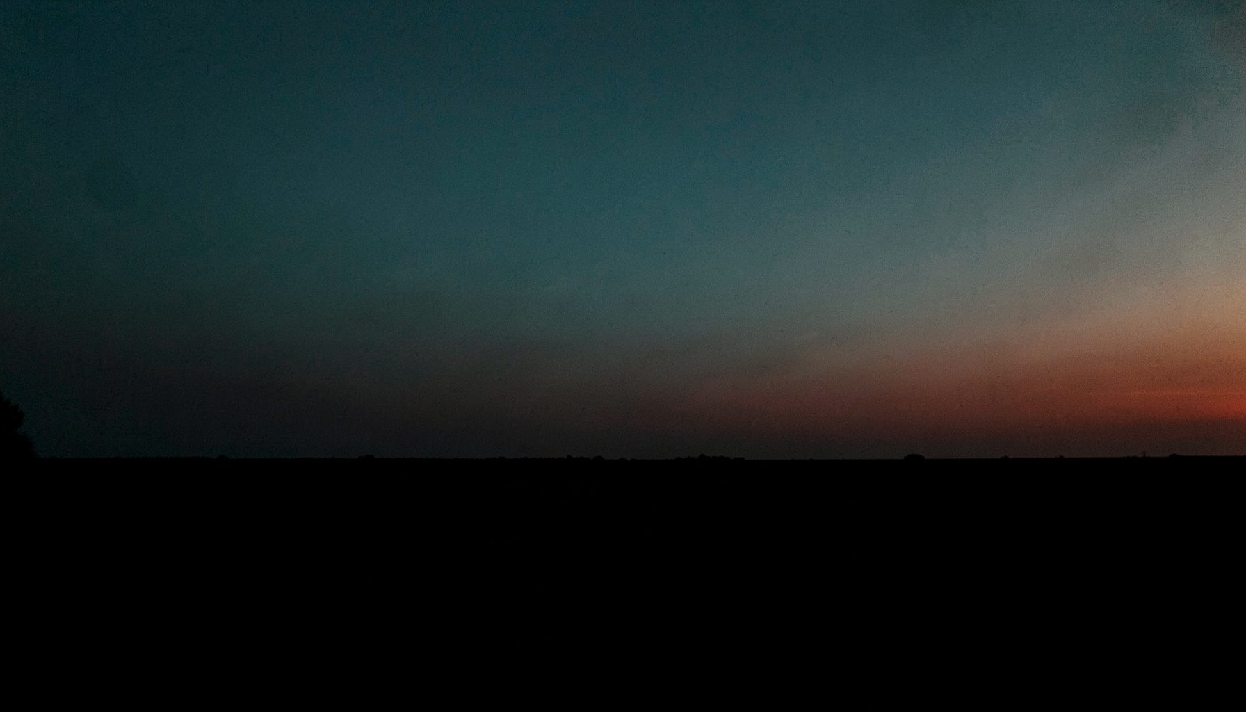Moody, low-key photograph of a long horizon at twilight, near-black foreground, muted teal sky, and a faint coral-red glow near the horizon